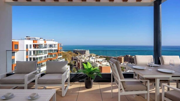 Une terrasse offrant une vue sur la mer, aménagée avec des chaises et une table blanches, ainsi qu’une plante en pot.