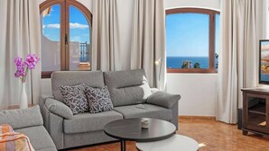 Un salon moderne avec un canapé gris, une table basse ronde et une vue sur la mer grâce à de grandes fenêtres.