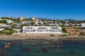 Ein Küstenhotel mit einem langen, weißen Gebäude, einem felsigen Strand davor und einer gebirgigen Kulisse.