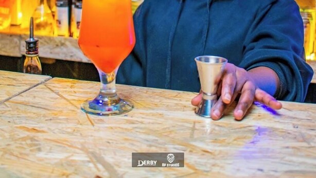 Un espace bar où l’on voit une personne tenant un shaker à cocktails, un grand verre rempli d’une boisson orange, et, au fond, des étagères remplies de bouteilles.