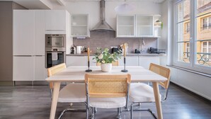 Een moderne keuken met witte kasten, een inbouwoven en een eethoek met een witte tafel en twee rieten stoelen.