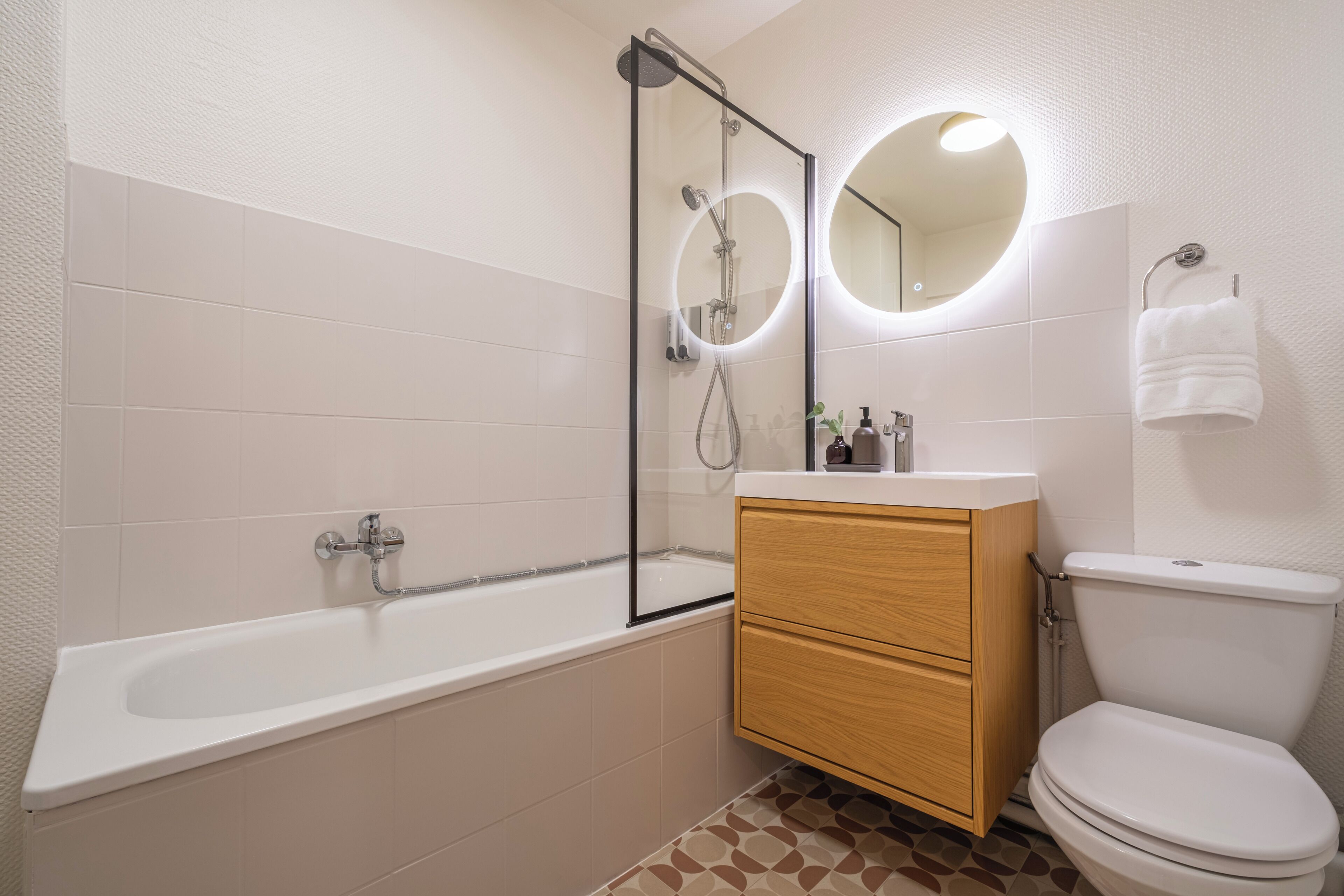 Une salle de bain moderne avec une baignoire, des toilettes, un meuble-lavabo avec des tiroirs et un grand miroir rond doté de lumières.