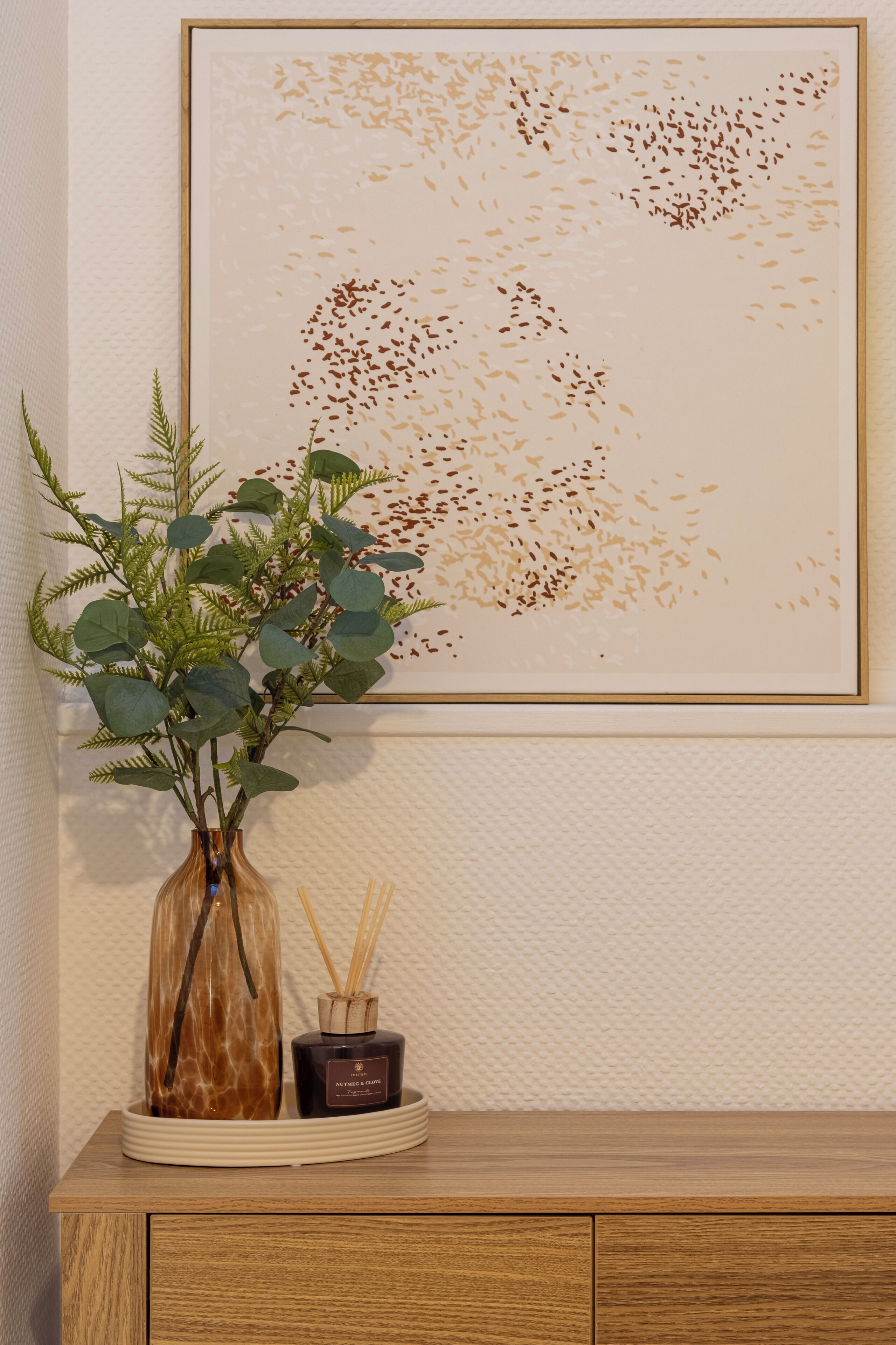 Een lijst met kunstwerk aan een muur, een houten kast met lades, een vaas met een plant en een klein potje met rietverspreiders.