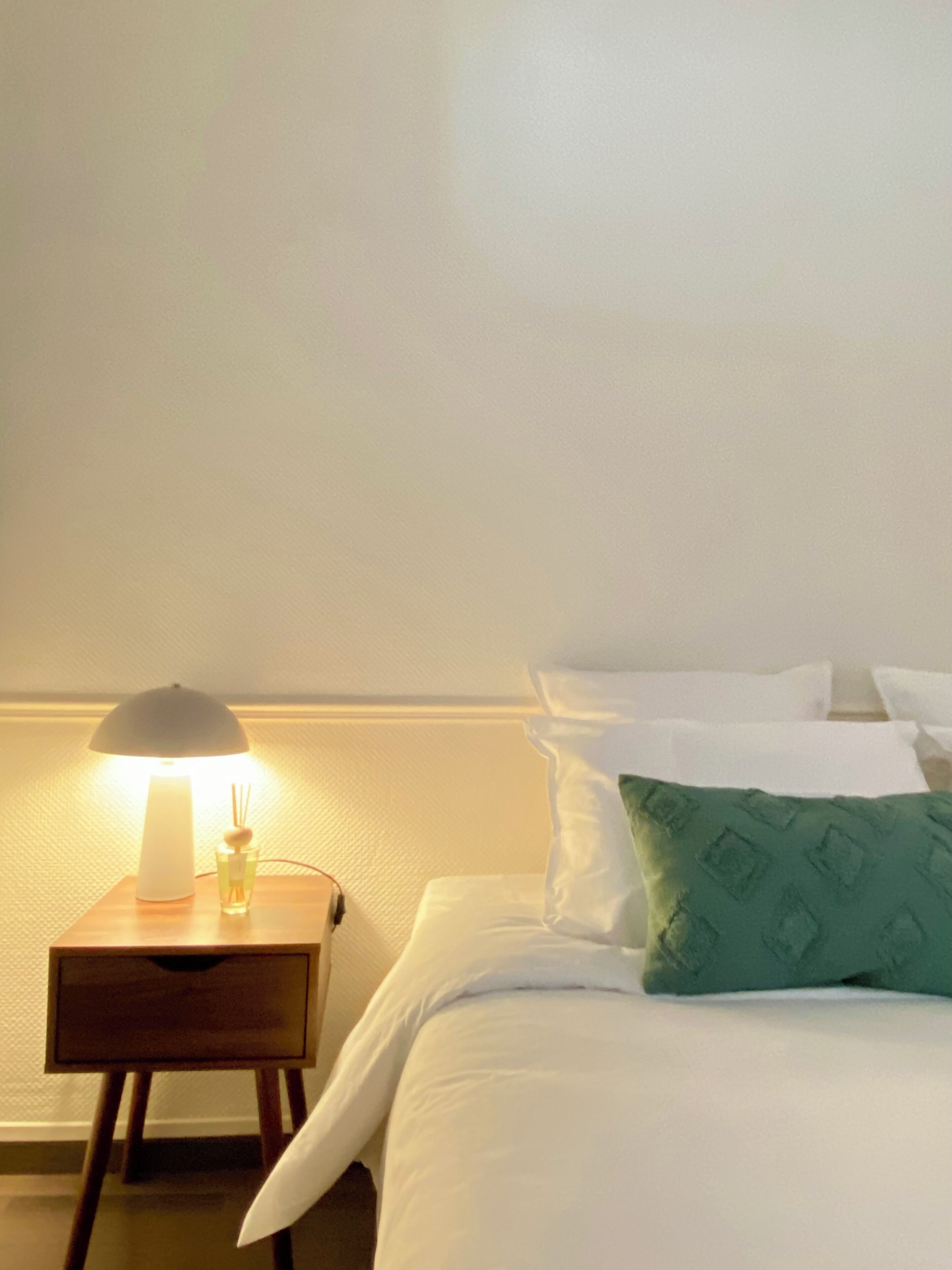 A neatly made bed with white linens and a green pillow, a bedside table with a lamp, and a small glass of water.