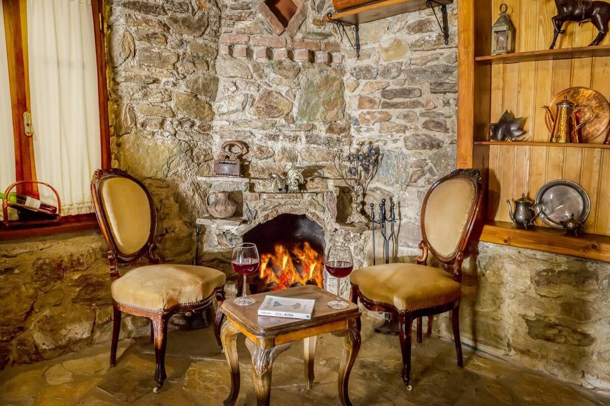 A stone fireplace with two chairs and a table in front, a shelf with decorative items, and a wooden shelf above the fireplace.