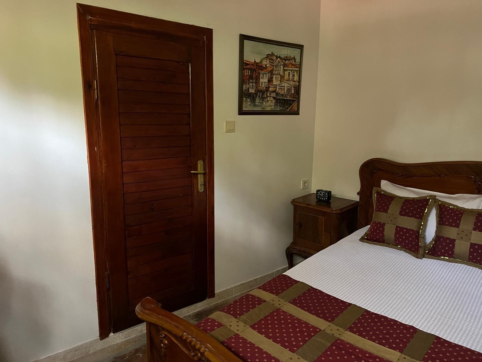 A bedroom with a wooden door, a bed with a quilt, a nightstand with a clock, and a framed picture on the wall.