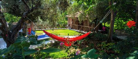 Una hamaca colgada entre árboles en un jardín con un pequeño edificio de ladrillo y una piscina azul al fondo.