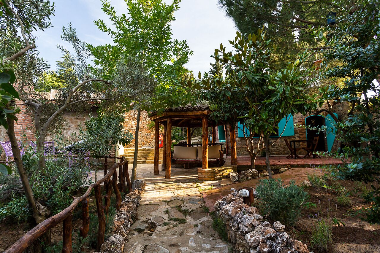 Un área de estar al aire libre rústica, con un pérgola de madera, un sendero de piedra y abundante vegetación.