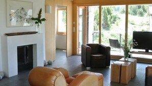 A spacious living area with a fireplace, leather sofa, and a large window offering a view of greenery. - La Montanella 4 by Interhome (Maloja)