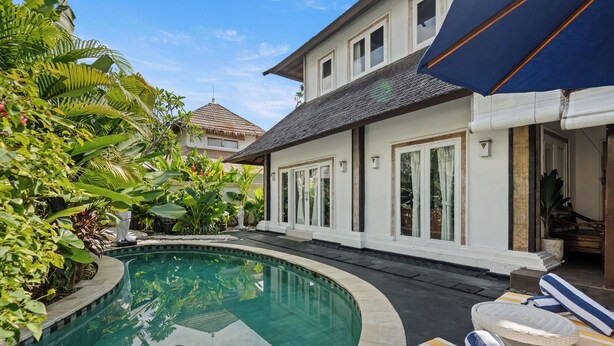 A swimming pool with lounge chairs and umbrellas, surrounded by lush greenery and a white building with large windows. - Canggu Wave Villa (Canggu)