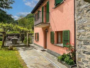 Un edificio rosa con contraventanas verdes, un muro de piedra y un jardín con pérgola.