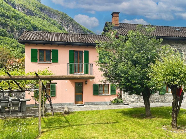 Una casa de dos pisos con un balcón, rodeada de exuberante vegetación y montañas.