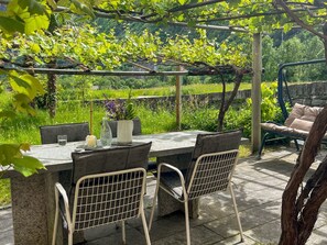 Un patio exterior con mesa y sillas, rodeado de follaje y una pérgola.