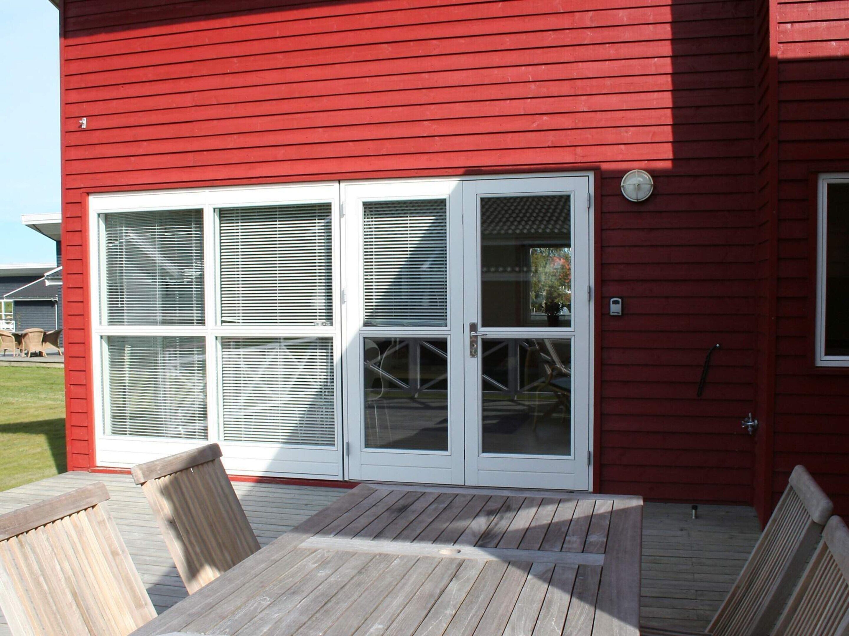 A red building with a large sliding glass door and a wooden deck with chairs.