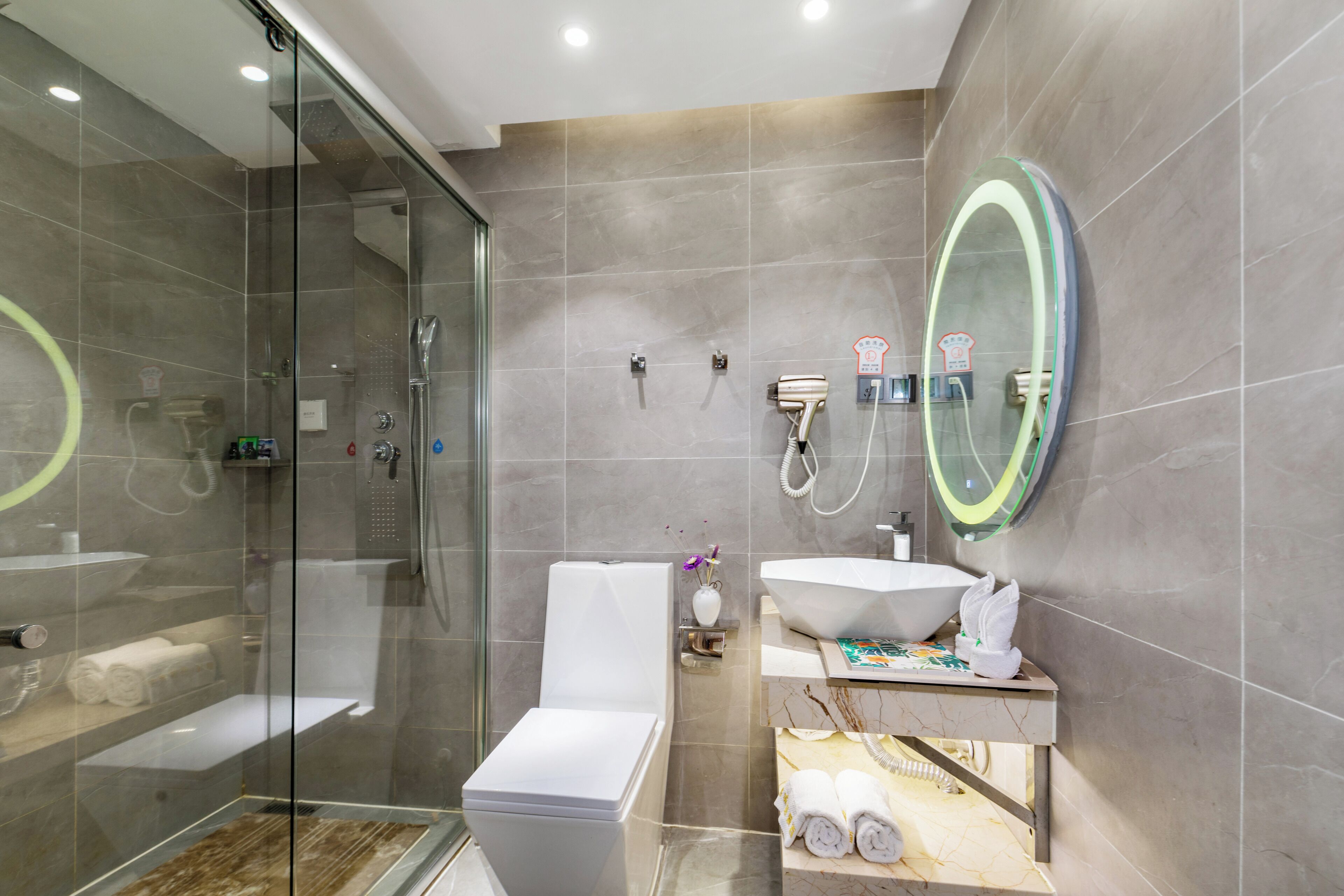 Un baño moderno con ducha de vidrio, inodoro blanco, un espejo ovalado grande y un mueble de tocador con lavamanos y toallero.