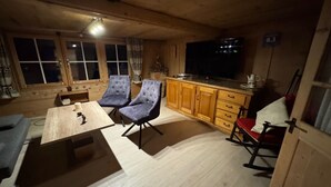 Tunnelmallinen mökin sisustus, jossa on puinen ruokapöytä, kaksi verhoiltua tuolia, keittiöalue, jossa on televisio, ja keinutuoli.