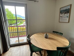 Ein Essbereich mit einem runden Holztisch, grünen Stühlen und Blick auf einen Balkon mit Geländer und umliegender Bepflanzung.