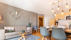 Ein modernes Wohnzimmer mit einer Couch, einem Glastisch und zwei Sesseln. Es gibt eine Küchenzeile mit Holzschränken und einem Esstisch.