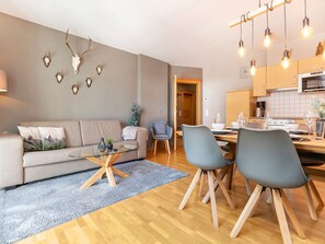 Ein modernes Wohnzimmer mit einer Couch, einem Glastisch und zwei Sesseln. Es gibt eine Küchenzeile mit Holzschränken und einem Esstisch.