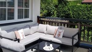 Een terras met een houten pergola, een witte bank met kussens, een klein tafeltje met een wijnglas, en uitzicht op een bebost gebied.