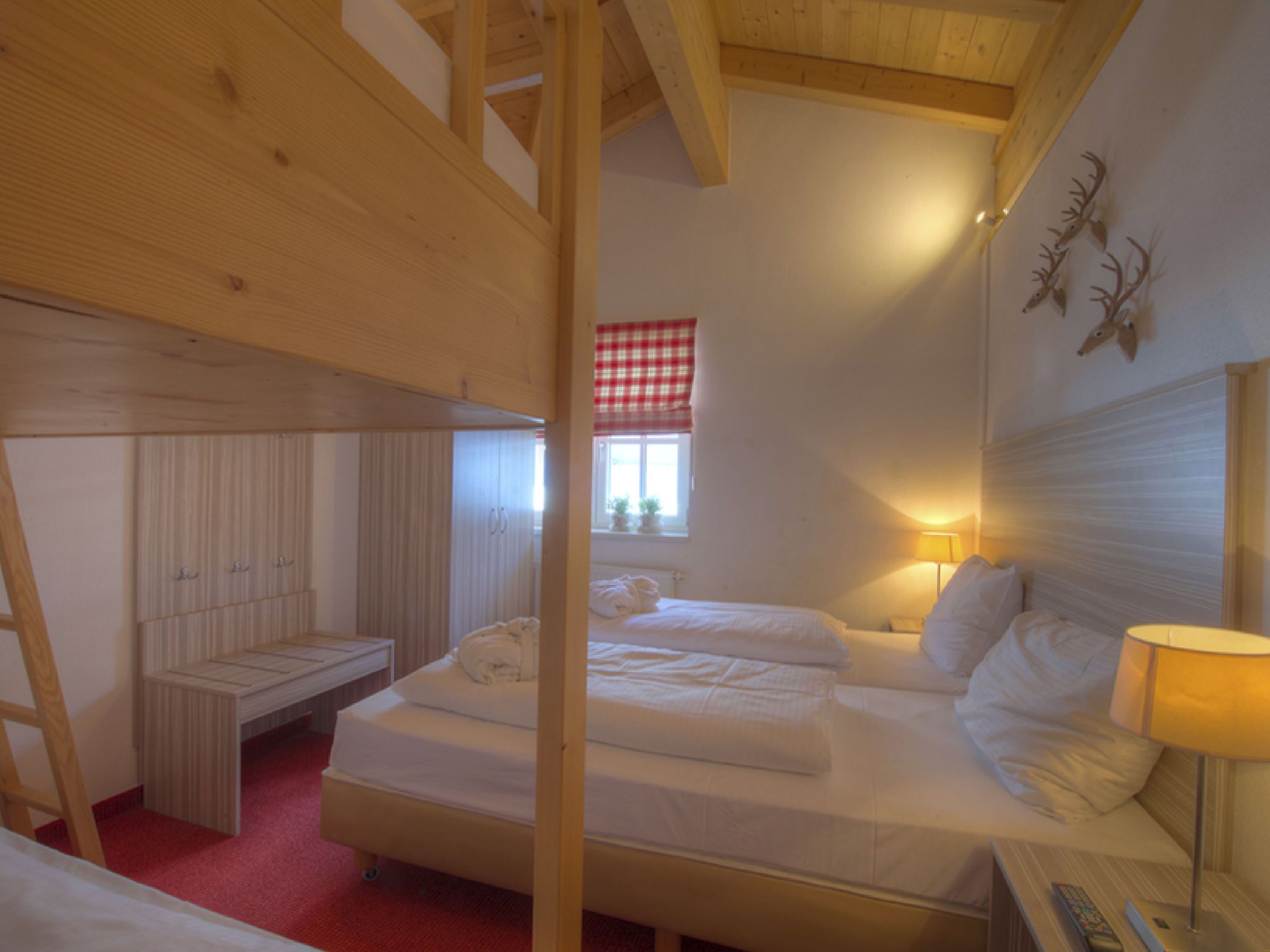 Una habitación con una cama individual superpuesta, una mesita de noche con lámpara, una ventana con cortina a cuadros rojos y una pared decorada con cornamentas de ciervo.