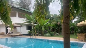 Une piscine dont le bord est revêtu de carrelage, entourée de verdure et bordée d’un bâtiment doté d’un balcon.