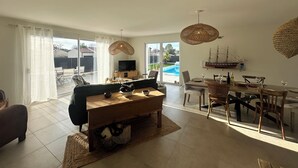Un salón amplio con un sofá grande, una mesa de centro de madera y un comedor con mesa y sillas. Hay un televisor sobre un mueble, un modelo de barco en la pared, y vistas a una piscina a través de puertas de cristal.