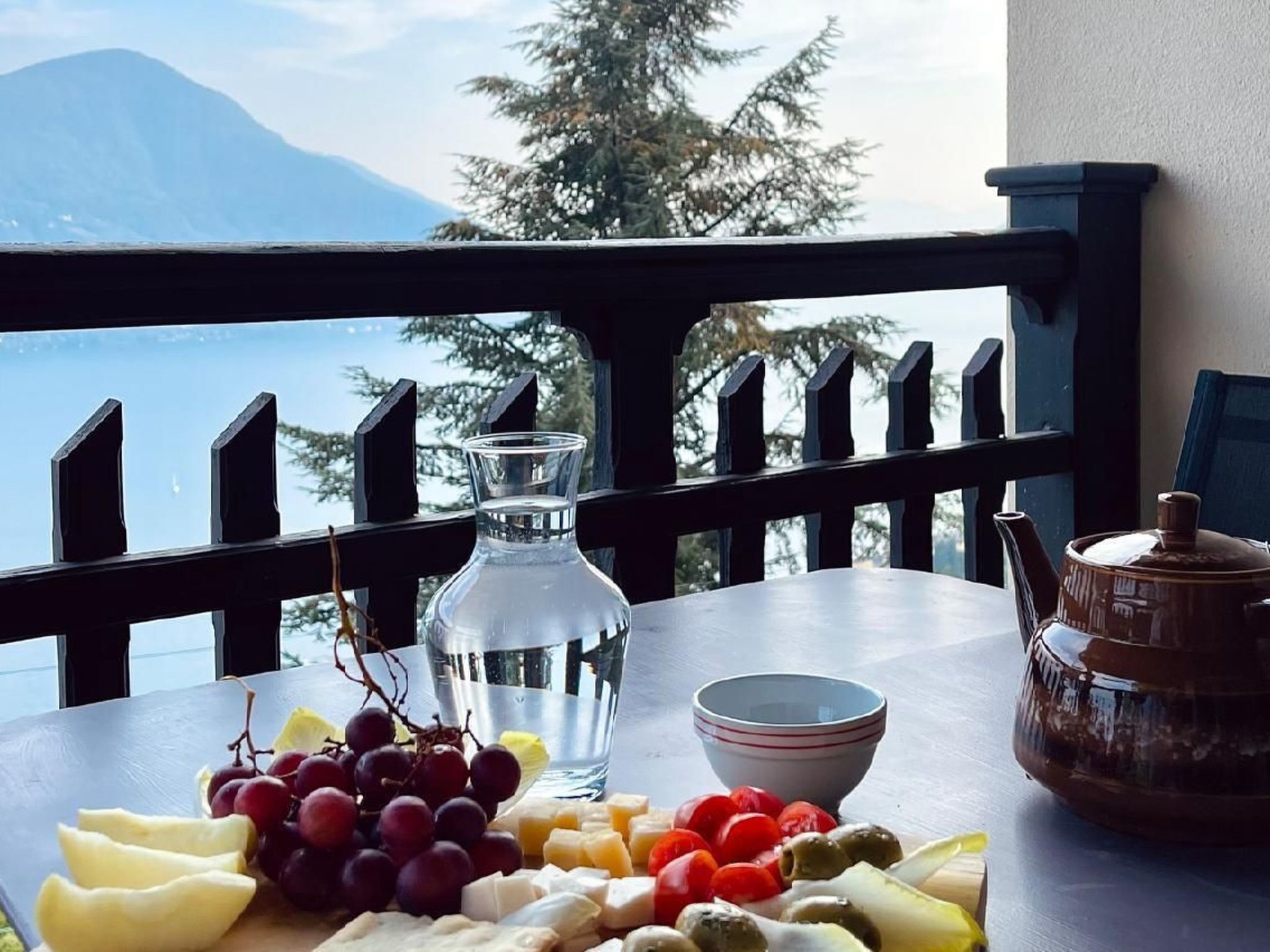 A table with a variety of fruits and cheeses, a glass pitcher, and a teapot on a balcony with a mountain view.