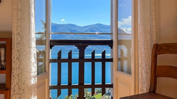 A room with a view of mountains and a lake through a window with a wooden railing. - Chalet Bazzi by Interhome (Brissago)