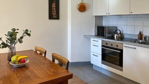 A modern kitchen with white cabinets, a microwave, kettle, and a sink. There's a dining table with chairs and a bowl of fruit. - Chalet Bazzi by Interhome (Brissago)