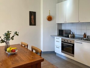 Eine moderne Küche mit weißen Schränken, einer Mikrowelle, einem Wasserkocher und einem Spülbecken. Ein Esstisch mit Stühlen und eine Schüssel mit Obst sind ebenfalls vorhanden.