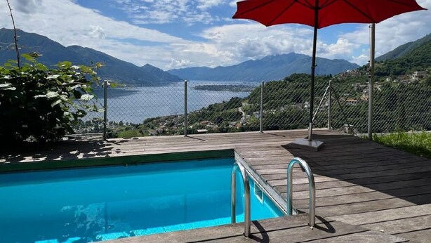 Eine Holzterrasse mit Swimmingpool, einem roten Sonnenschirm und Blick auf Berge und einen See.