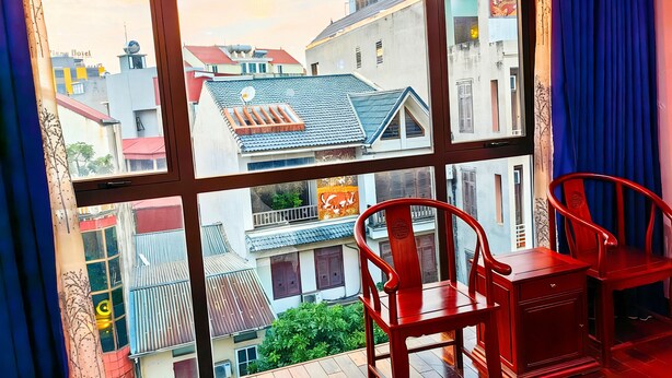 A room with a large window offering a view of neighboring buildings and a balcony with red chairs and a wooden cabinet. - HEYI HOTEL (Tu Son)