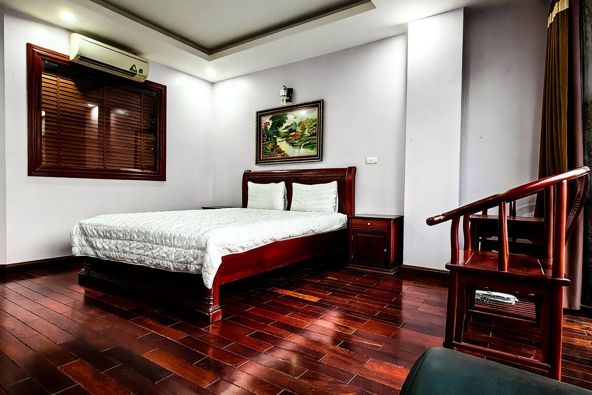 A bedroom with a wooden bed, a nightstand, a wall-mounted air conditioner, a framed painting, and a staircase.