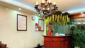 A reception area with a marble floor, a wooden reception desk, a chandelier, a clock, and a wall-mounted menu board. - HEYI HOTEL (Tu Son)