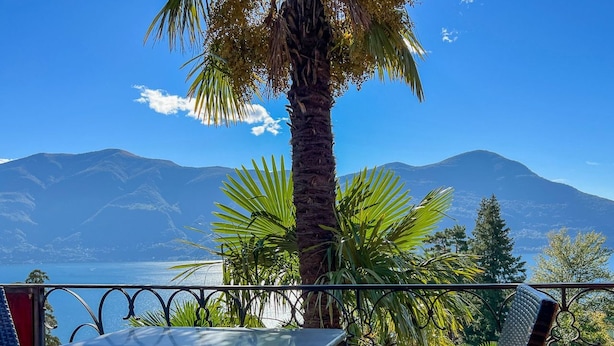 Varanda com mesa e cadeiras, vista para um corpo de água e montanhas.