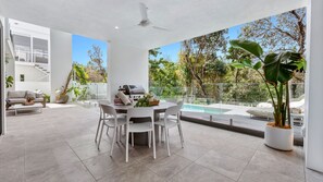 A modern outdoor dining area with a pool, a ceiling fan, and a view of the surrounding greenery.