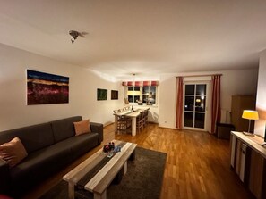 Ein Wohnzimmer mit einer Couch, einem Couchtisch und einem Essbereich mit Tisch und Stühlen. Ein Fenster mit Vorhängen und eine Tür zu einem anderen Raum sind ebenfalls vorhanden.