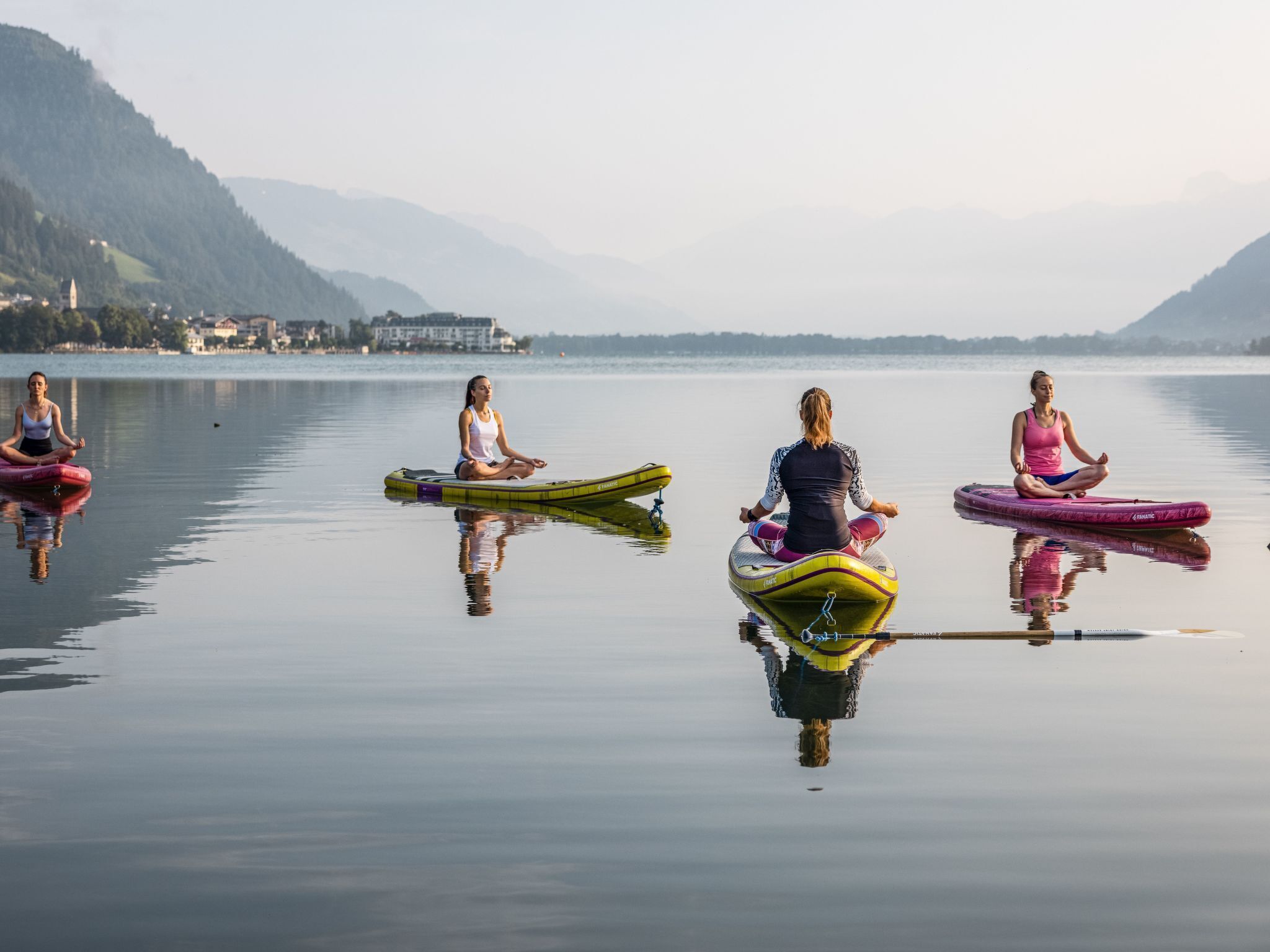 Cuatro personas meditando en botes inflables individuales sobre un lago tranquilo, con montañas al fondo.