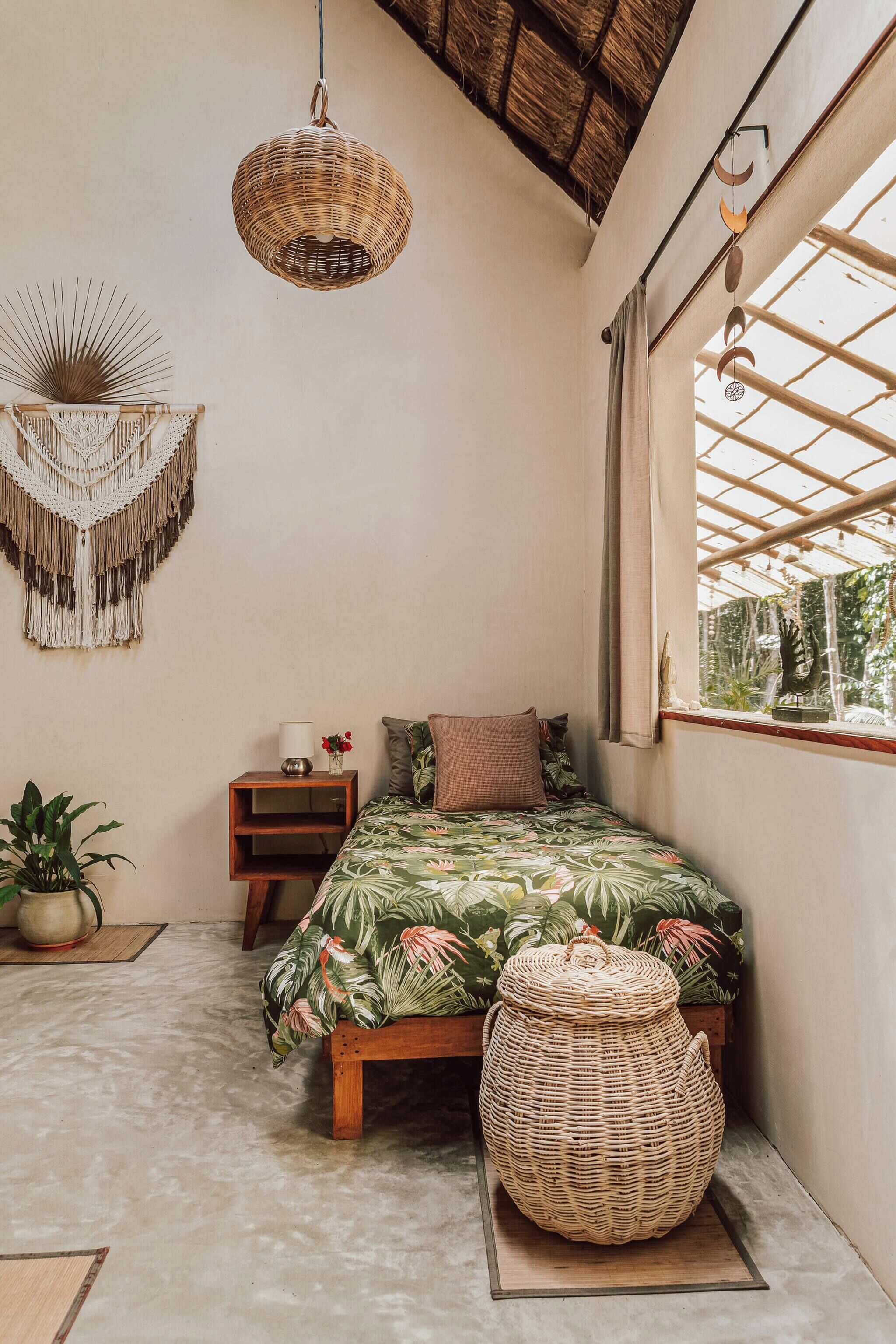 A bedroom with a bed, bedside table, hanging light, macrame wall decor, and a window with a view of greenery.