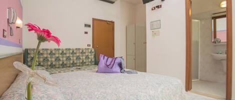 A bedroom with a patterned bedspread, a purple pillow, a vase with a pink flower, a wooden door, and a bathroom visible through an open door.