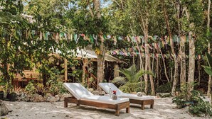 Two wooden lounge chairs with white cushions on a sandy surface, surrounded by lush greenery and a string of colorful flags. - Room-casa Arkaana Jungle Ecocasita Share (Tulum)