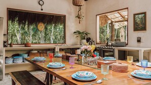 A dining area with a long wooden table set for a meal, surrounded by chairs. There are various dishes, glasses, and a vase with flowers on the table. - Room-casa Arkaana Jungle Ecocasita Share (Tulum)