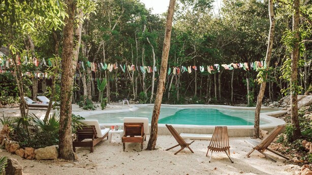 A poolside area with a sandy beach, wooden lounge chairs, and a tropical setting with trees and a hanging string of flags. - Room-casa Arkaana Jungle Ecocasita Share (Tulum)