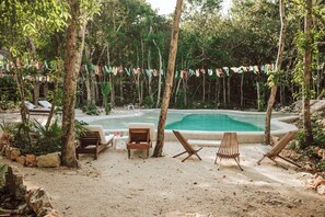 Ein Poolbereich mit Sandstrand, Holzliegen und tropischer Umgebung mit Bäumen und einer girlandenförmig hängenden Flaggenreihe.