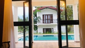 Une vue à travers une porte vitrée donnant sur un espace piscine, avec un bâtiment blanc en arrière-plan.
