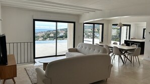 Ein großzügiger Wohnbereich mit einer Couch, einem Esstisch und Stühlen, große Schiebeglastüren zu einem Balkon mit Blick auf das Meer und Gebäude.