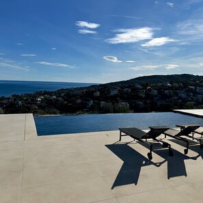 Ein Pool ohne Ende mit Liegestühlen, blauem Himmel und Blick auf eine Küstenstadt.