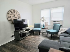A living room with a flat-screen TV, a large wall clock, two armchairs, a sofa, and a window with blinds. - Gorgeous Updated 2-Bedroom Cottage Near Mount Mary University & Mayfair Mall (Milwaukee)
