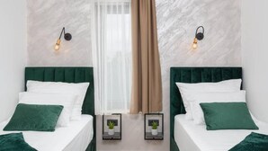 Habitación doble con dos camas, cada una con cabeceros verdes y ropa de cama blanca. Hay luces fijadas en la pared a cada lado de un ventanal con cortinas traslúcidas.
